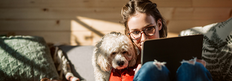 A woman working on a tablet computer while cuddling a dog, in a home setting.