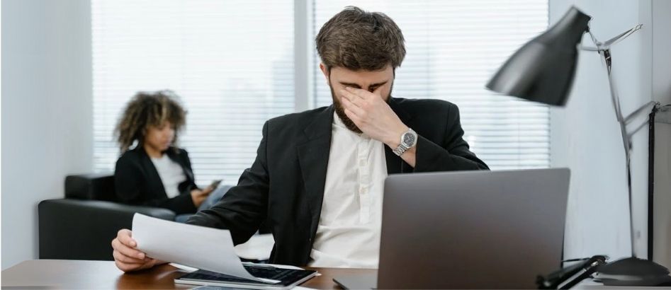 A frustrated man in a suit sits at a desk with a laptop, engaged in a tense salary increment discussion.