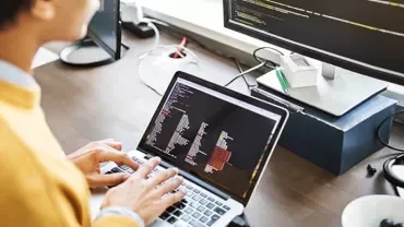 Person using a laptop and external monitor with code editor open.