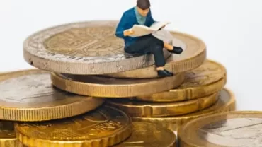 A miniature figure reading a newspaper on a stack of coins.