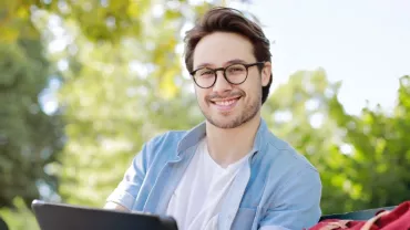 One Caucasian man in casual clothes holding a laptop and sitting on a garden bench and smiling to camera