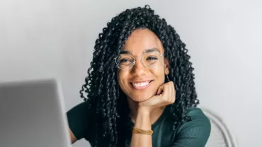 A person smiling while sitting in front of a laptop wearing glasses.
