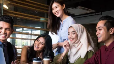 Five friends smiling while looking at a laptop in a café.