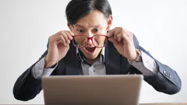 An Asian, white-collar professional holding out his glasses for a better look at his laptop screen