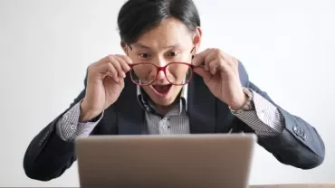 Person looking at a laptop screen, holding glasses with a surprised expression.