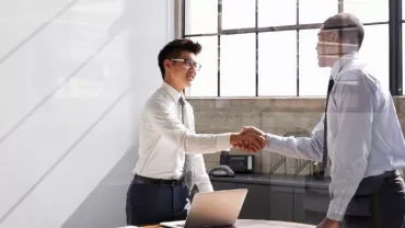 Two people shaking hands in an office with sunlight streaming through the windows.