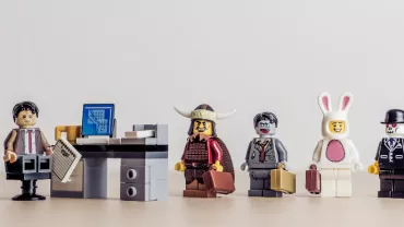 Five LEGO figures stand by a desk, each uniquely dressed, holding briefcases.