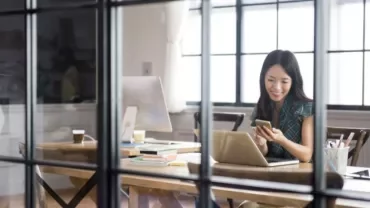 Person using phone and laptop in office, with books and coffee on the desk.