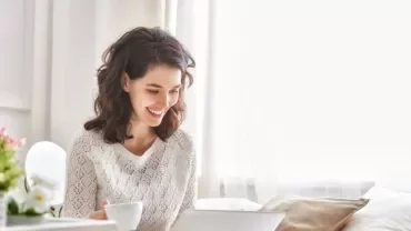 Person smiling while using a laptop in a bright room.