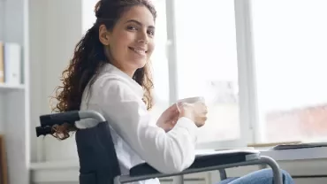 A person sitting in a wheelchair holding a cup, smiling near a laptop.