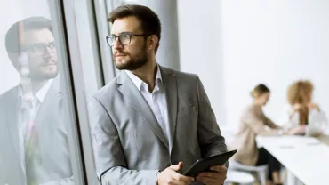 A person in a suit looks out a window, with colleagues in a meeting in the background.