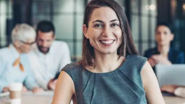 Person smiling in an office, with colleagues in the background having a discussion.