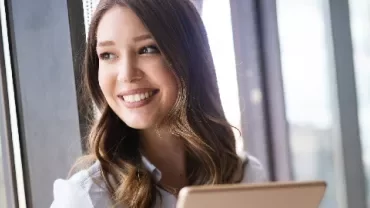 Person smiling while holding a tablet by a window.