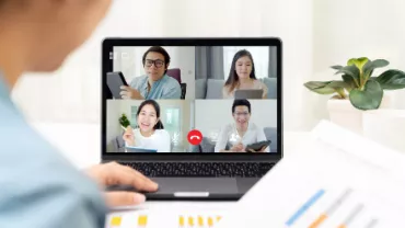 Person in a virtual meeting with three others on a laptop screen, papers and a plant in the background.