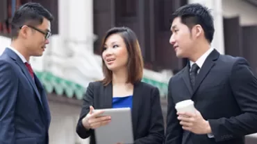 Three people standing outdoors having a conversation; one holds a tablet.