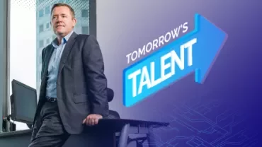 Person in a suit standing by a desk with 'Tomorrow's Talent' sign in the background