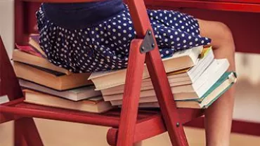 A child wearing a blue polka dot dress sits on a stack of books.