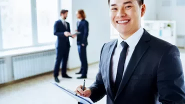 Person in a suit smiling and holding a clipboard in an office setting.