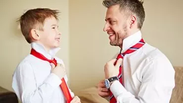 An adult teaches a child to tie a tie, both smiling.