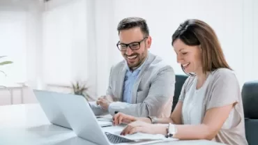 辦公室中的兩個人在使用筆記本電腦，面帶笑容。