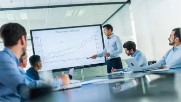 A meeting with a speaker presenting a chart, while others listen.