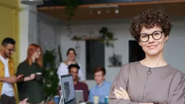 Person with glasses smiling in an office, with a team meeting in the background.
