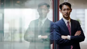 A person in a suit leaning on a glass wall with folded arms.