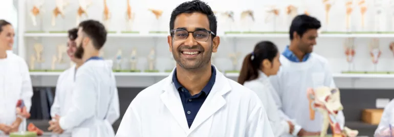 A person in a white lab coat smiling in a lab with colleagues and anatomical models in the background.