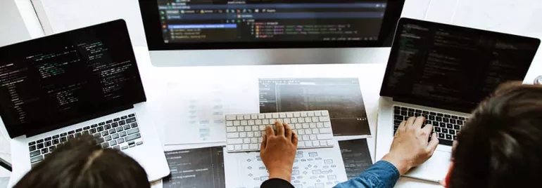 Several people coding on laptops with a large monitor displaying code.