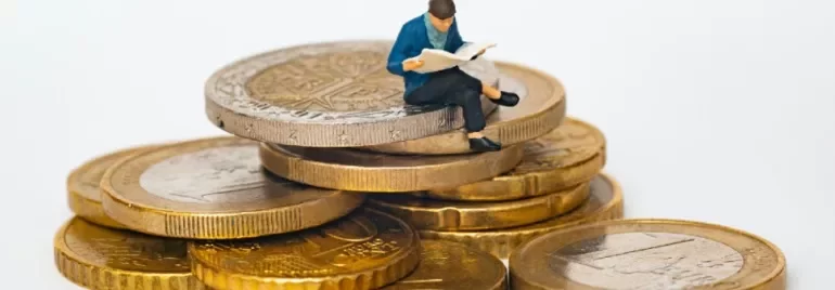 A miniature figure reading a newspaper on a stack of coins.