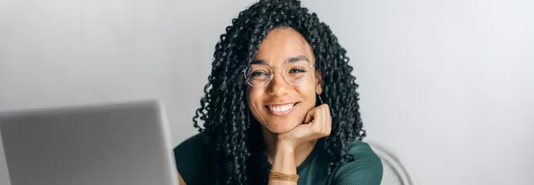 A person smiling while sitting in front of a laptop wearing glasses.