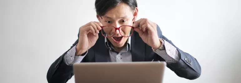 Person looking at a laptop screen, holding glasses with a surprised expression.