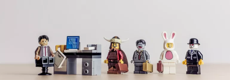 Five LEGO figures stand by a desk, each uniquely dressed, holding briefcases.
