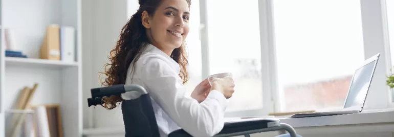 A person sitting in a wheelchair holding a cup, smiling near a laptop.