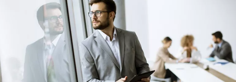 A person in a suit looks out a window, with colleagues in a meeting in the background.