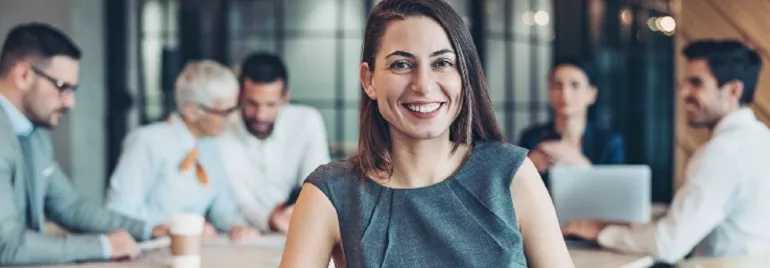 Person smiling in an office, with colleagues in the background having a discussion.