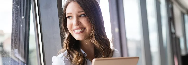 Person smiling while holding a tablet by a window.