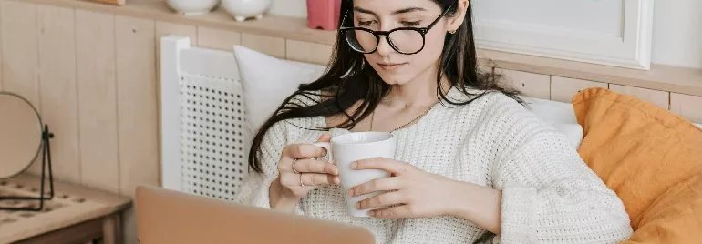 Person on a couch sipping coffee while using a laptop.