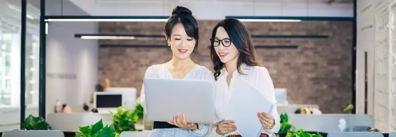 Two women in an office collaborate over a laptop, discussing career growth opportunities and talent retention strategies.