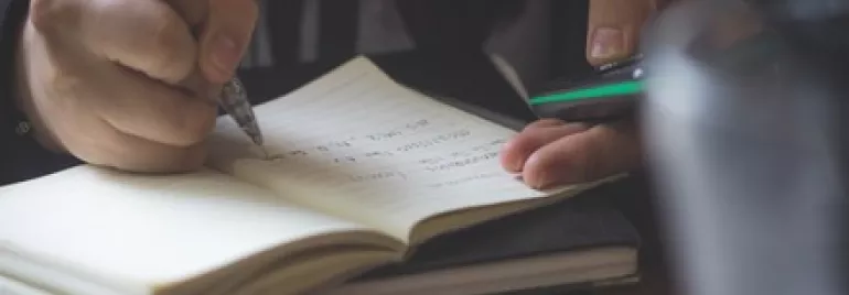 一雙手正在筆記本上書寫，另一隻手拿著手機。