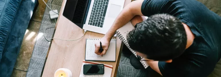 Overhead view of a person writing in a notebook at a table with a laptop and phone.