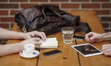 Two people taking notes at a coffee table with a notebook and tablet.