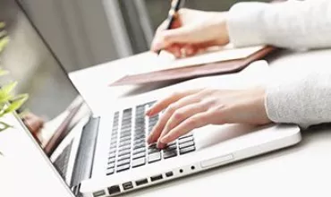 A person typing on a laptop while writing in a notebook.