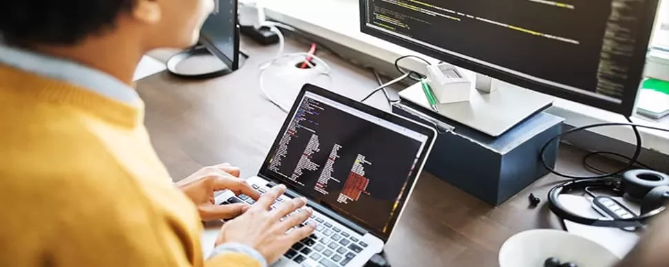 Person using a laptop and external monitor with code editor open.