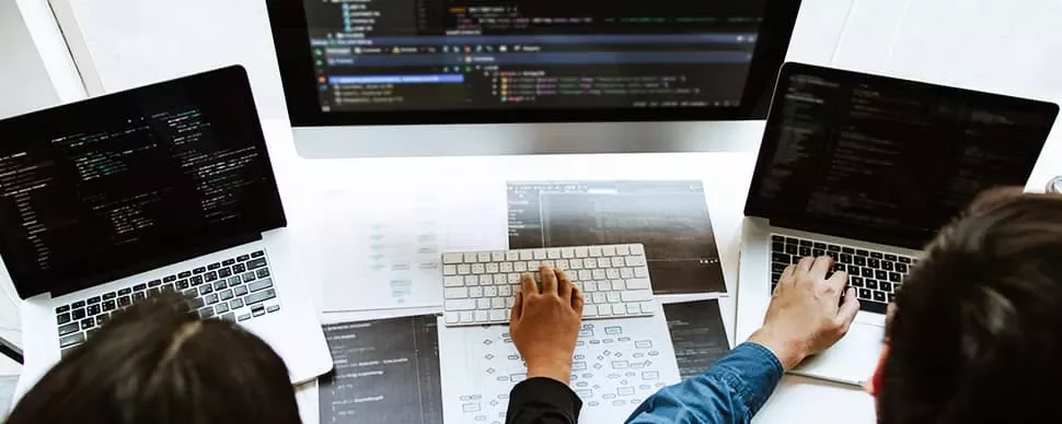 Several people coding on laptops with a large monitor displaying code.