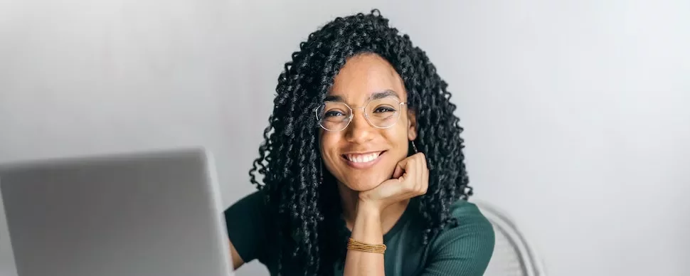 A person smiling while sitting in front of a laptop wearing glasses.