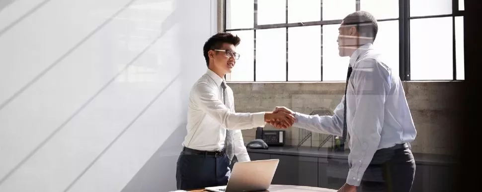 Two people shaking hands in an office with sunlight streaming through the windows.