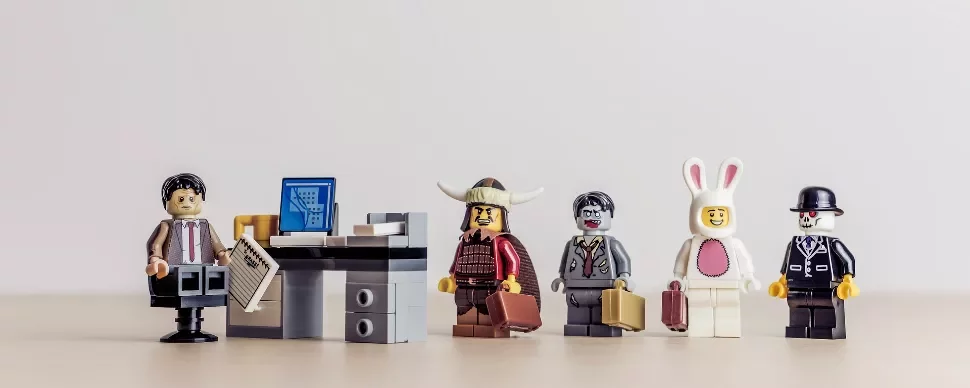 Five LEGO figures stand by a desk, each uniquely dressed, holding briefcases.