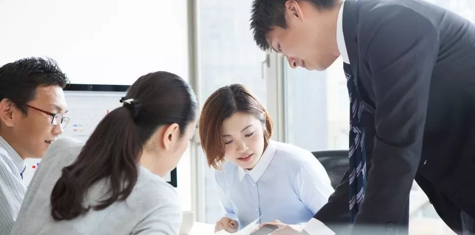 Group of people engaged in discussion at an office desk with a computer screen in the background.