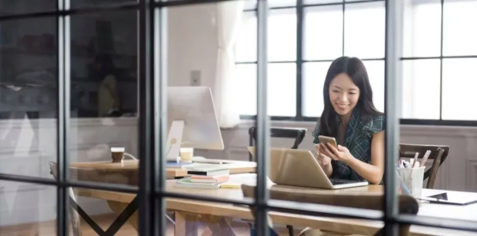 Person using phone and laptop in office, with books and coffee on the desk.